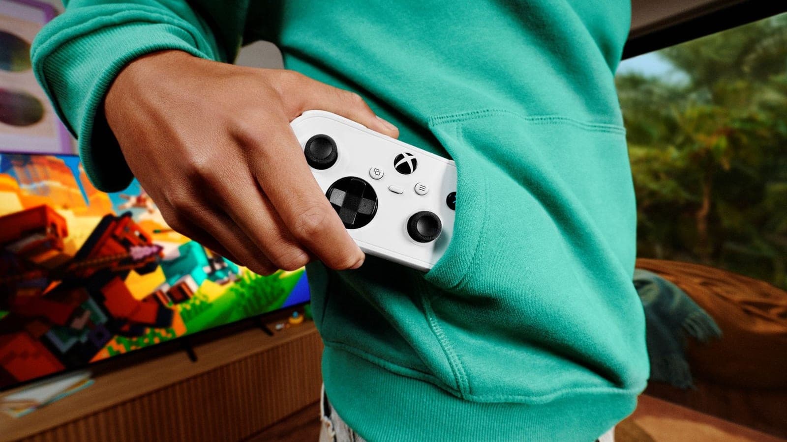 A person in a green hoodie slips a small white Xbox Nano controller into the front pocket, with a Minecraft game on a TV behind them